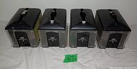 Four stainless steel paper napkin dispensers with black tops shown in a row, front facing, on white tablecloth with green number card.