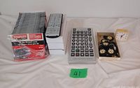 Photo showing slim CD/DVD cases stacked, four regular size universal remote controls stacked on a white box, one jumbo universal remote control with large buttons, ten decorative keychains in a white box, and a small beige frame clock.