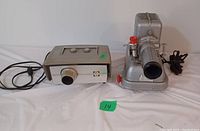 Two vintage slide projectors on a white background, one grey rectangular and one metallic silver with red knob.