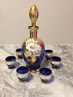 Full set of one cobalt blue Bavarian decanter with gold and floral design and six matching shot glasses arranged around it on a grey surface.