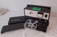 Photo showing the Kodak 8mm projector unit with metal film reel mounted and black carrying case next to it on a white surface