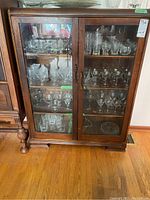 Front view of vintage wood cabinet filled with glassware, showing two glass paneled doors, metal handles, and wood base with minor scratches and wear.
