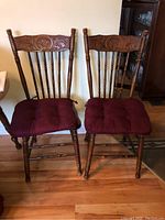 Two wooden chairs side by side showing carved backrests with ornate floral and scroll designs and burgundy padded cushions on seats.