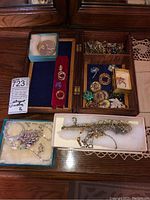 Open wooden jewelry box showing compartments filled with brooches, rings, purse hook box, gold-tone chains, crystal necklace set