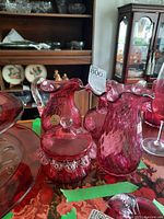Table display showing cranberry glass candy bowl with lid and hanging drops, cranberry glass vase, and cranberry glass pitcher with clear handle and original label