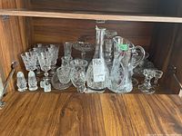 Full view of crystal glassware assortment on wooden shelf, includes pitchers, decanters, bowl, candlesticks, salt & pepper shakers, cream and sugar set, and glasses.