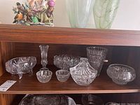 Shelf display of eight clear crystal glass items including various candy dishes, bowls, and vases with cut and etched patterns.