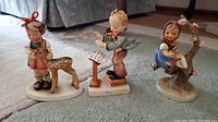 Front view of three Hummel figurines showing girl with deer, boy conductor, and girl on tree stump.
