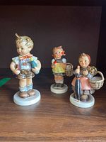 Three Hummel figurines on a shelf: boy in lederhosen, girl with basket, girl with umbrella and basket.