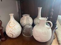 Three white Kaiser vases and one white jug arranged on a wooden shelf, showing intricate raised and gold details.