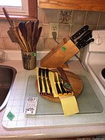 Photo of wooden knife block with knives, wooden spoons in a ceramic holder, set of steak knives on decorative cloth on round wooden board, and glass cutting boards on counter