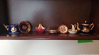 Shelf containing seven Limoges porcelain decorative pieces with gold accents including teapots, plates, basket, and lidded container.