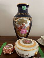 Front view of large Limoges vase with black background, gold detailing, and romantic couple scene, alongside a round trinket box and larger jewelry box