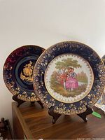 Two 10-inch Limoges plates on wooden stands, one with a blue and gold intricate border and couple scene, the other with a gold figure on dark background.