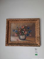 Framed painting on canvas showing pink and blue flowers in a vase with an ornate gold frame, minor crazing visible on canvas.