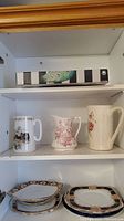 Shelf with three decorative pitchers (pastoral, red floral, pink rose) and sets of decorative plates beneath.