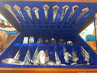 Open wooden cutlery box with blue velvet lining holding assorted forks, spoons and serving pieces
