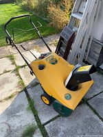 Front side angled view showing yellow body, black chute and handle with controls, four wheels.