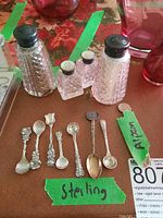 Image showing salt and pepper shakers with polished sterling silver lids and 8 miniature spoons, including 1 marked Alpaca. All items on brown surface with floral background.