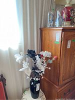 Black vase with faux white and dark flowers displayed on a small round table in front of a window