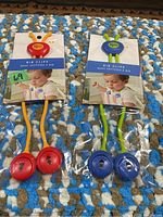 Two pairs of Kipris bib clips in packaging: one red with orange arms, one blue with green arms, placed on a multicolored carpet