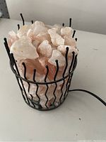 Photo shows a Himalayan salt lamp with multiple irregular pink salt chunks inside a black metal wire basket. The basket has a zigzag pattern and the lamp has a visible power cord.