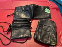 Black genuine leather small crossbody bag and fold-over wallet laid flat on red surface. The bag has multiple zippered compartments and a black adjustable string strap. The wallet has a smooth texture with fold-over closure.