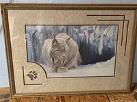 Framed print of a bobcat in snow with tree and icicle background. Wood frame with paw print cutout in mat.