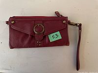 Front view of the red leather wallet displaying flap with decorative ring buckle and snap closure along with attached wrist strap and zippered compartment.