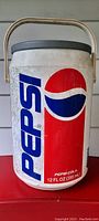 Front view of a white hard plastic cooler with the large vintage Pepsi logo in red, white, and blue. Cooler has a black lid and a metal carry handle.