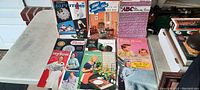 Photo of vintage sewing and craft magazines and books with colorful illustrated covers showing vintage women sewers, crocheters, and craft projects including dolls, embroidery, and home decor ideas.