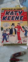Photo shows front cover of a Katy Keene comic book from the Archie series. Cover artwork features cartoon characters including a snowman wearing a tuxedo and a woman in red attire. The comic is old and shows signs of wear.