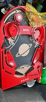 Top angled view of the red Brio countertop pinball machine showing space graphic playfield with planet and sun designs, flippers, metal ball guides, and plastic components. Bag with chrome pinball balls visible on top.