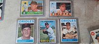 Photo of 5 vintage Baltimore Orioles baseball cards in protective sleeves on a beige surface. Cards show Jackie Brandt, Milt Pappas, Dave McNally, Luis Aparicio, and Johnny O'Brien.