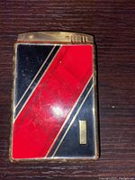 Front view of vintage Art Deco combination cigarette lighter and cigarette case showing red and black diagonal enamel panels separated by gold-tone lines on metal body.