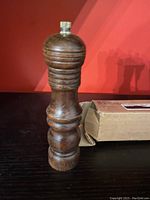 Front view of the wooden peppermill standing upright next to its shipping box against a red background