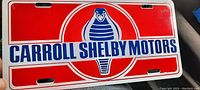Front view of vintage tin license plate for Carroll Shelby Motors with cobra logo and red, white, and blue colors.