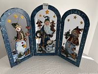 Front view of the three-panel metal Christmas fireplace guard with painted snowman and Santa designs and star decorations.
