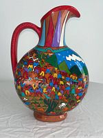 Front view of the round-bodied ceramic pitcher showing colorful folk art scene with people and animals in a village landscape with mountains in the background, red handle, and spout.