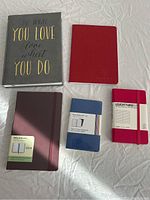 Five notebooks and planners laid flat showing different sizes and colors, including grey cover with gold text, red notebooks, and blue pack