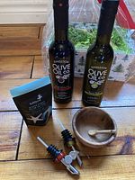 Full set front view showing 2 olive oil bottles, salt package, wooden bowl and spoon, and 2 pour spouts arranged on wooden floor.