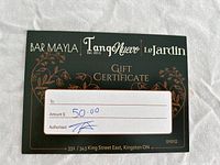 Front side of a black gift certificate card showing logos of Bar Mayla, Tango Nuevo, and Le Jardin. It includes a floral design, locations, and handwritten amount $50 with authorization.