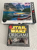 Photo showing a 1000-piece puzzle box with a vibrant image of canoes on a lake and a Star Wars Origami book with paper folding projects
