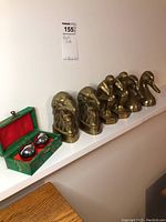 Six brass-look metal book ends and two shiny metal musical balls in green box with red lining on shelf against white wall.