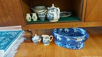 Various Churchill dishes and serving pieces including salt and pepper shakers, cream and sugar set, teapot, and a large ceramic serving pot. Items shown on wood floor and in cabinet.