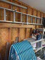Aluminum extension ladder hung horizontally on a wooden garage wall above shelves storing various household items