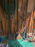 Photo showing the assortment of garden tools arranged standing against the wall including shovels, hoses, brooms, rake, and fork.