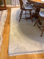 Close-up of the carpet under a dining table showing embossed scroll and floral design in light color with fringe edges.