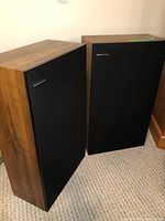 Pair of Boston Acoustics A70 speakers with wood veneer cases and black fabric front covers standing on carpeted floor.