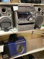 Front view of JVC MX-K5 stereo system showing central unit and two speakers on a shelf, with remote control and papers on top.